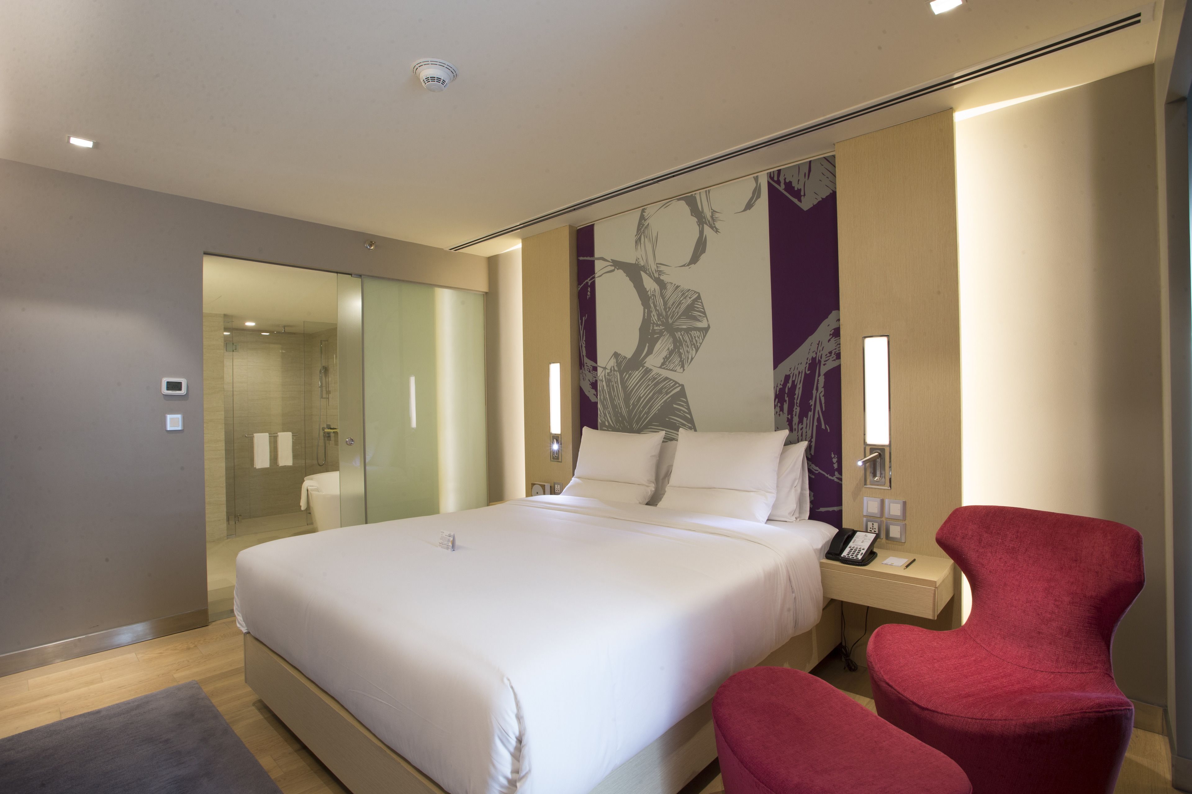 Una habitación de hotel moderna con una cama grande, un sillón rojo, una mesita de noche con un teléfono y un box de ducha de vidrio.