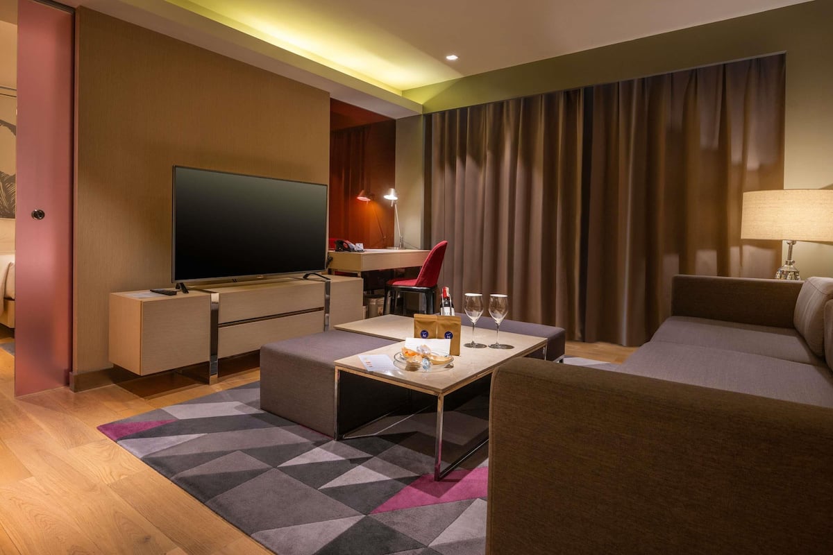 A modern hotel room with a flat-screen TV, a desk with a chair, a sofa, a coffee table, and a patterned rug.