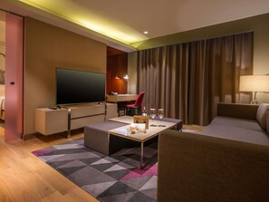 A modern hotel room with a flat-screen TV, a desk with a chair, a sofa, a coffee table, and a patterned rug. - Novotel Manila Araneta City Hotel (Quezon City)
