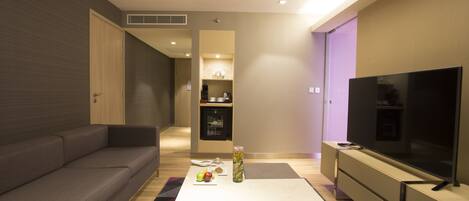 A modern living room with a sofa, a coffee table with fruits and a vase, a flat-screen TV, and a kitchen area in the background.