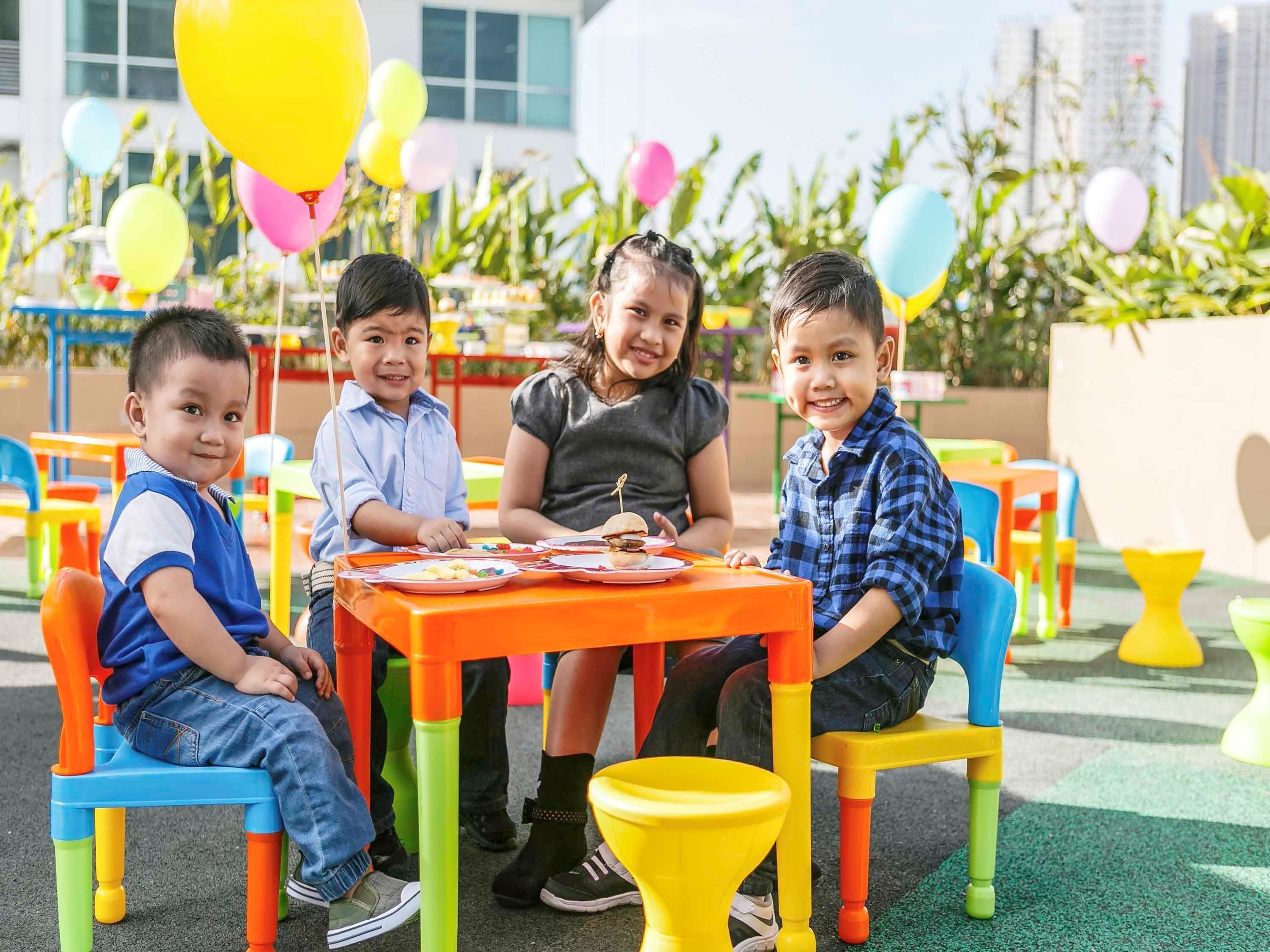 Children seated at colorful tables outdoors with balloons in the background.