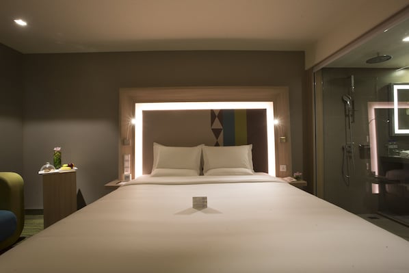 Una habitación de hotel moderna con una cama grande, una mesita de noche con lámpara, una pequeña mesita auxiliar con un jarrón, y una zona de ducha visible a través de una puerta de cristal.