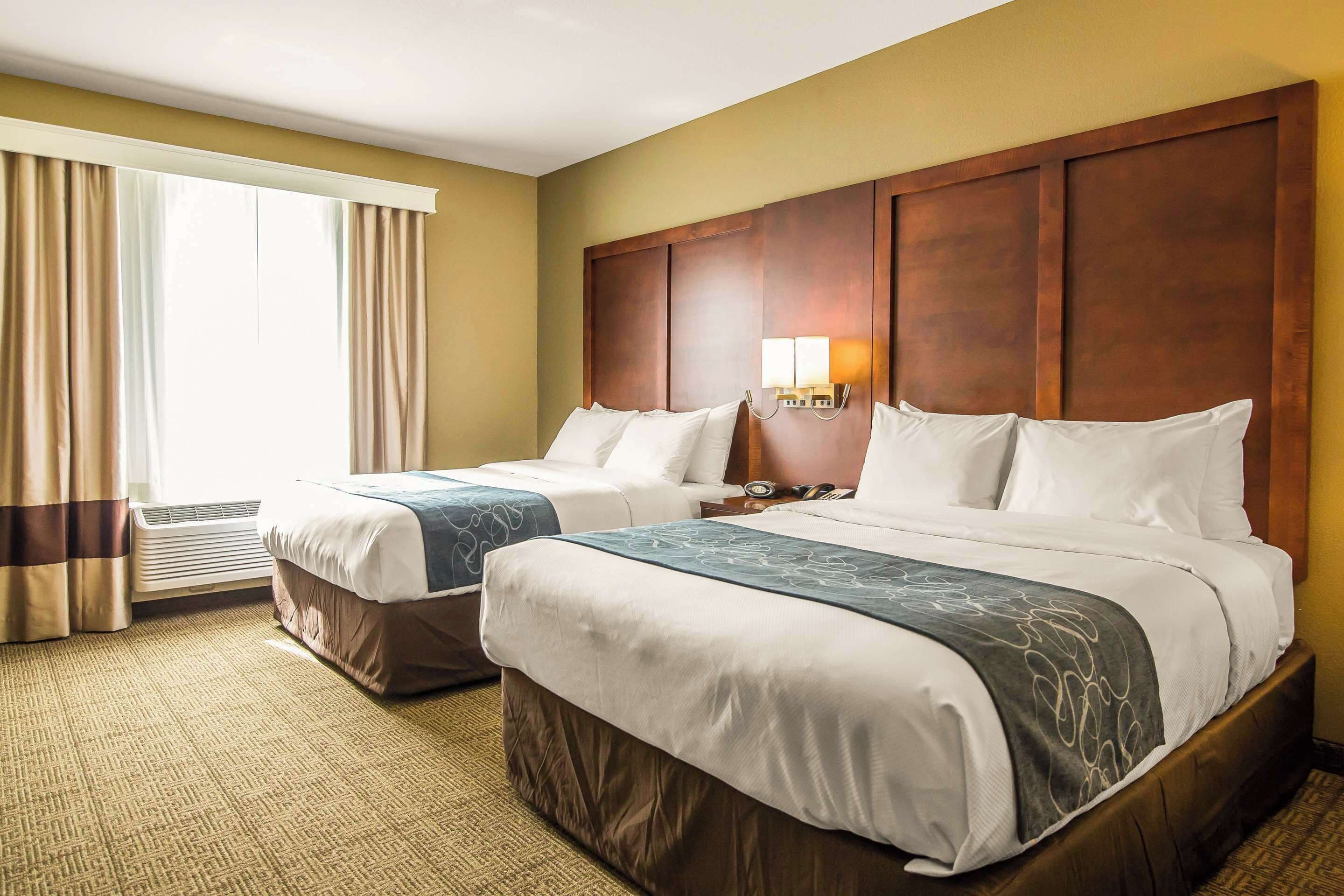 Habitación de hotel con dos camas, un ventanal con cortinas, cabecera de madera y una lámpara de pared.
