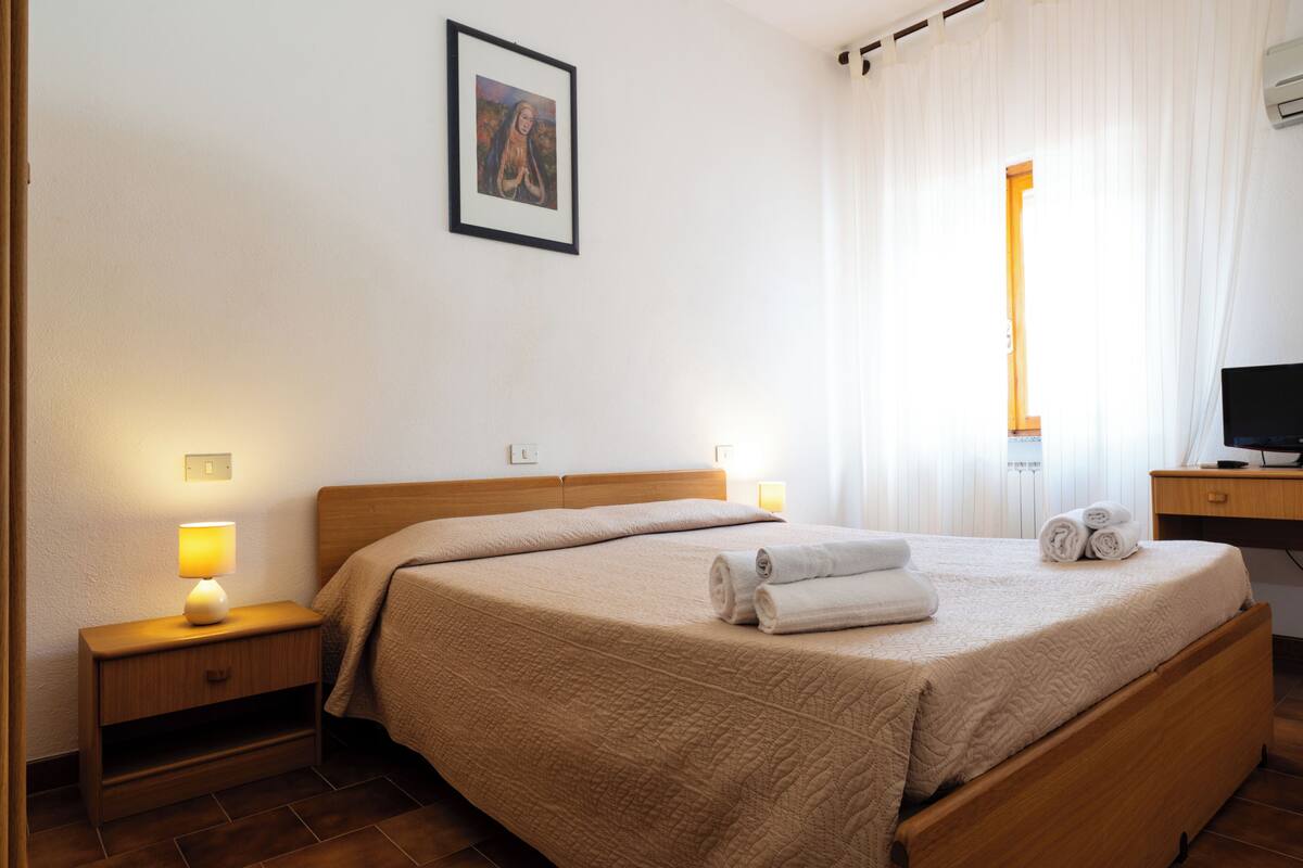 Un letto rifatto con ordinata precisione, dotato di una testiera, due comodini con lampade, un quadro incorniciato appeso al muro e una finestra con tende.