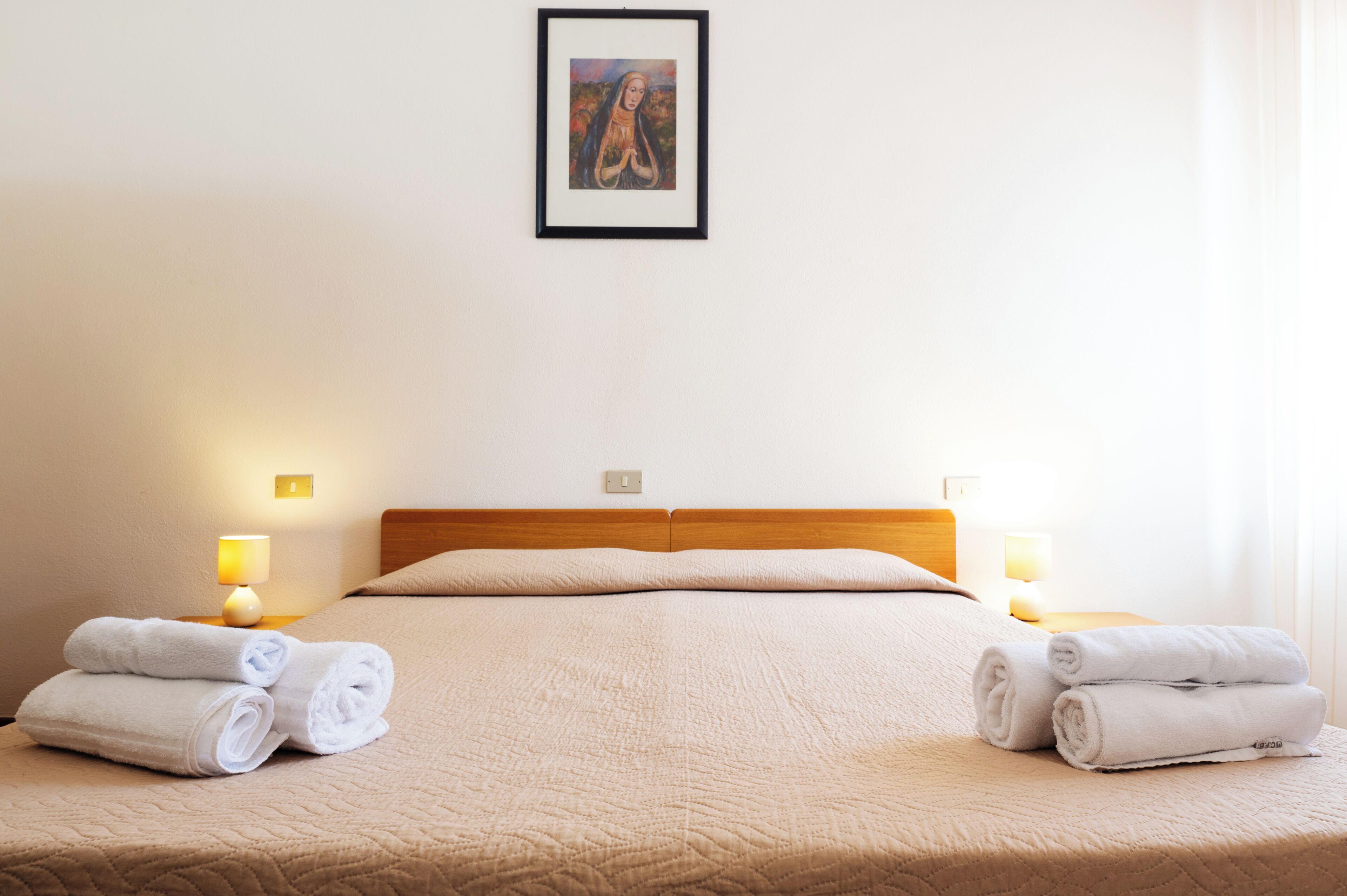 A neatly made bed with a beige quilt, two bedside lamps, and two rolled towels on the bed.