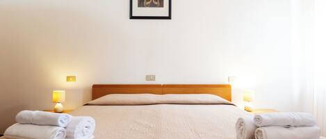 A neatly made bed with a beige quilt, two bedside lamps, and two rolled towels on the bed.