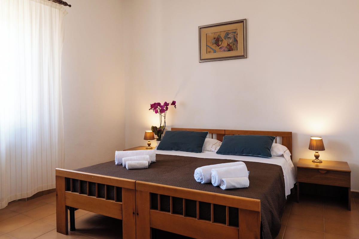 Camera d'albergo con un letto di legno, due comodini con lampade, un vaso con fiori e un quadro appeso al muro.