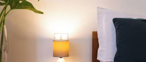 A hotel room with a bedside table, a lamp, a cup, a pillow, and a plant.