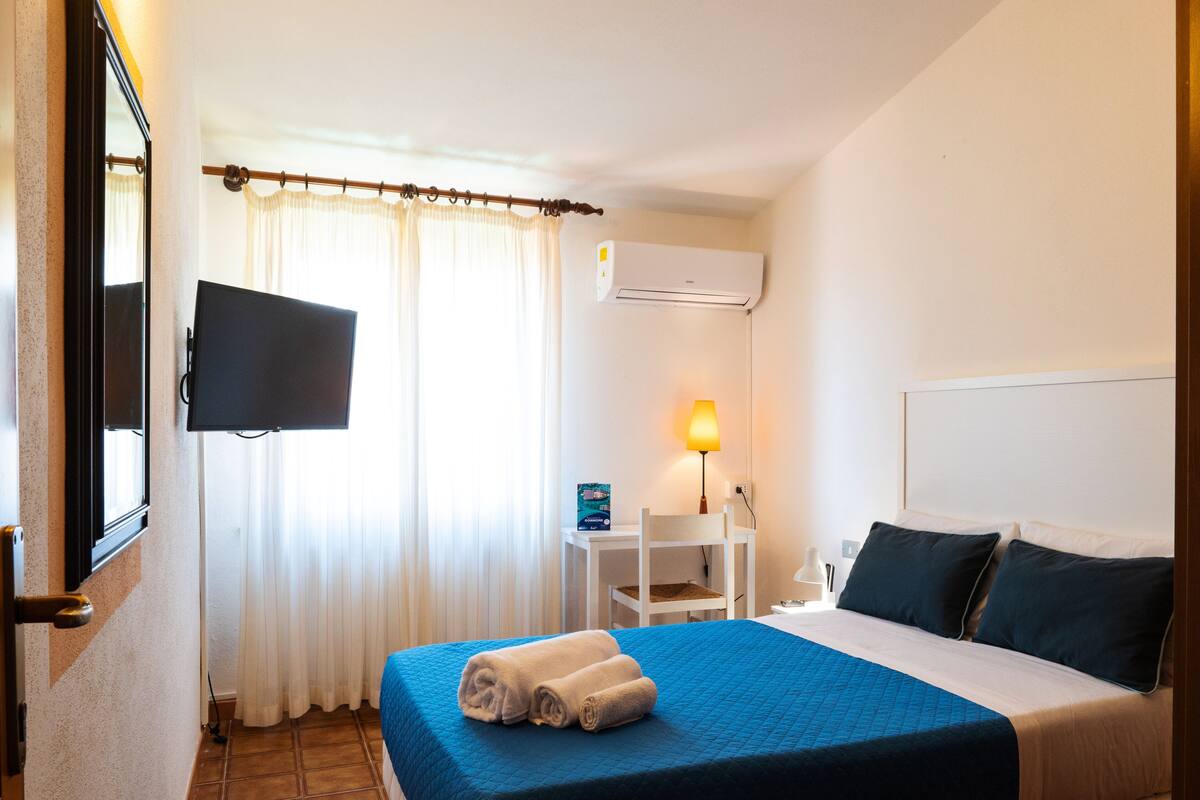 Una camera d'albergo con un letto, una TV a muro, una scrivania con una sedia, una lampada e un condizionatore d'aria.