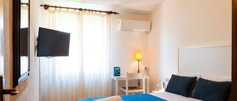A hotel room with a bed, a TV mounted on the wall, a desk with a chair, a lamp, and an air conditioning unit.
