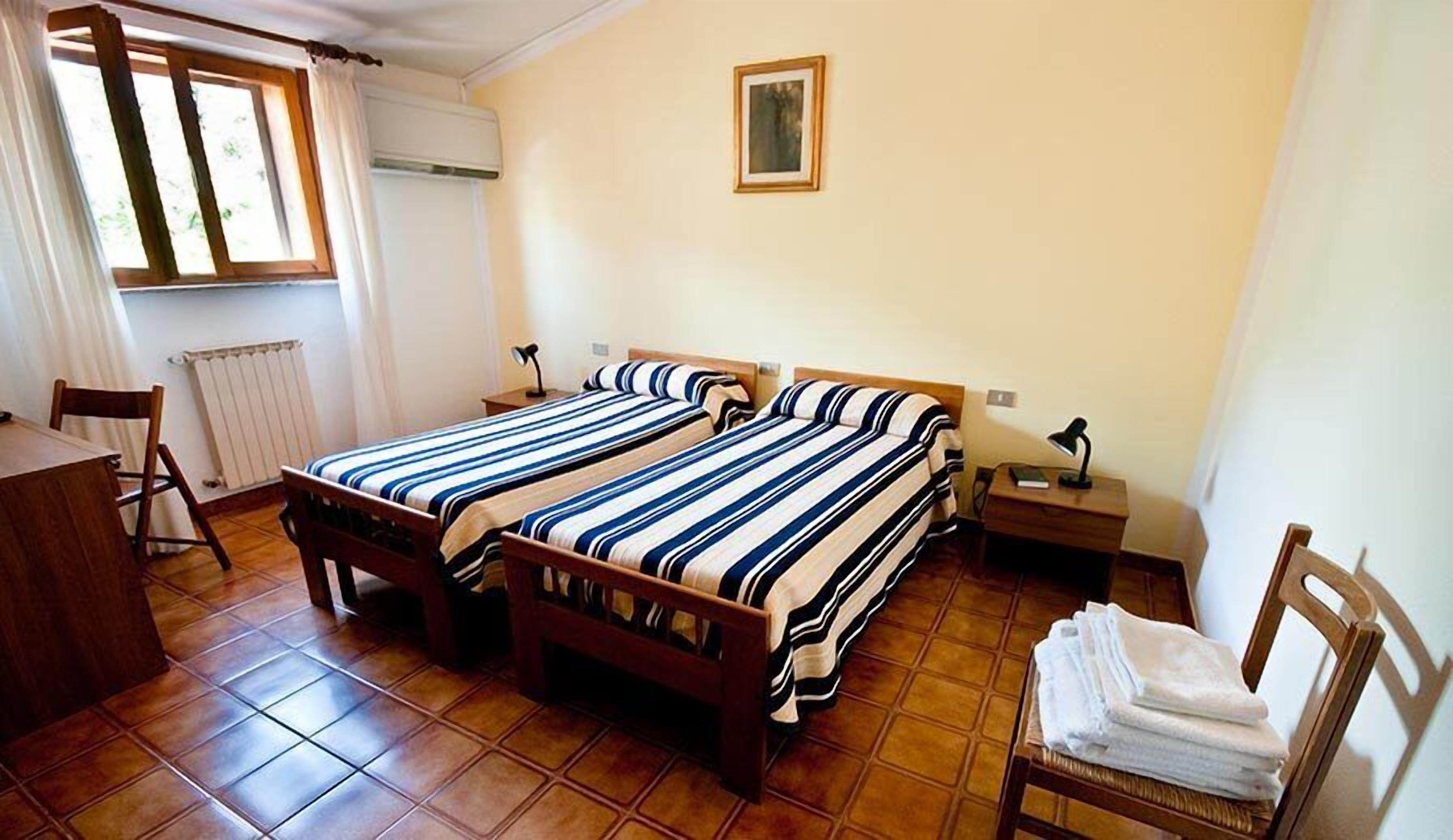 A room with two wooden beds, a desk, a chair, a radiator, a window with curtains, a nightstand with a lamp, and a framed picture on the wall.