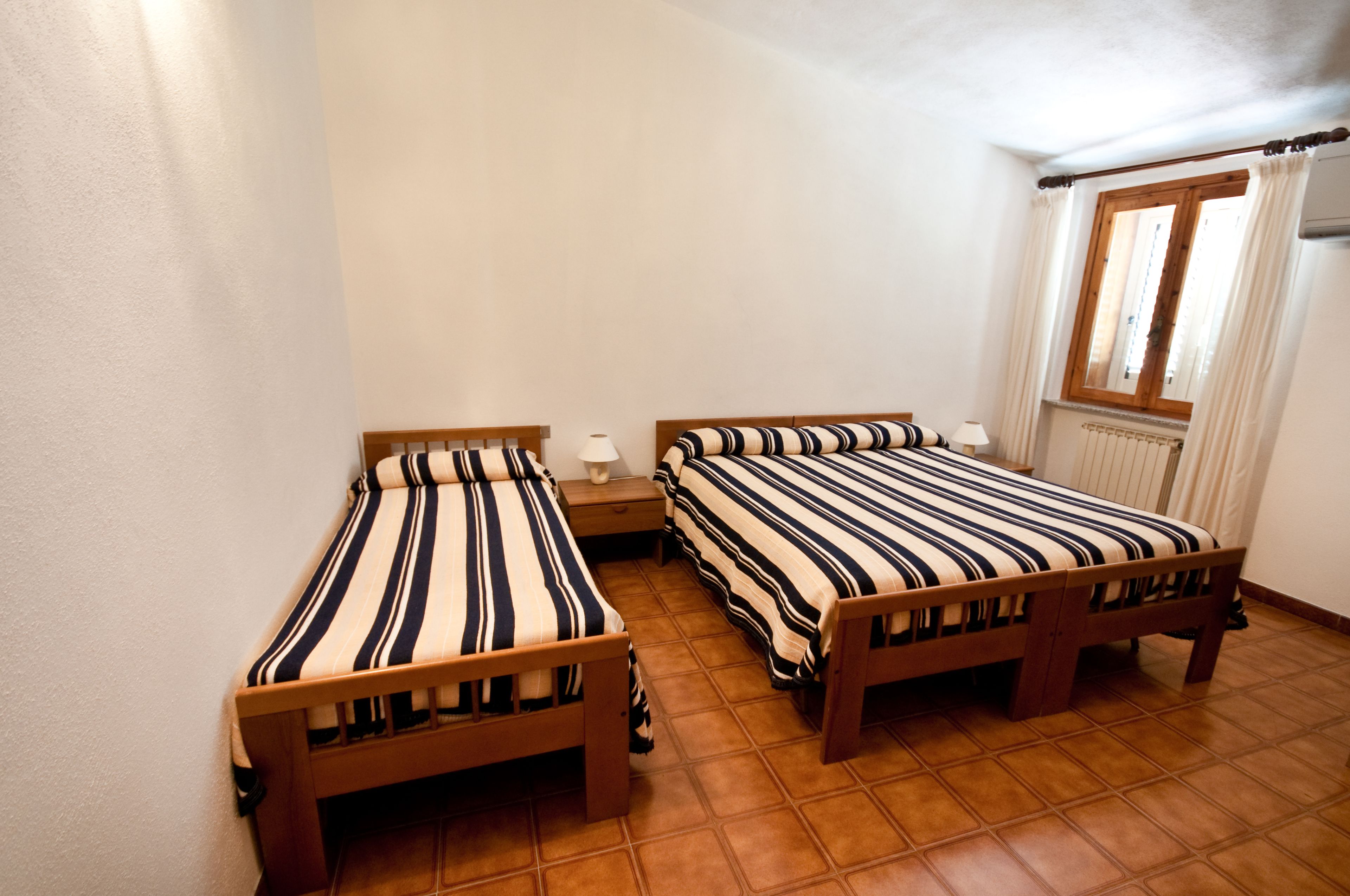 Ein kleines, sauberes Zimmer mit zwei Holzbett, einem Nachttisch und einem Fenster mit Vorhängen.