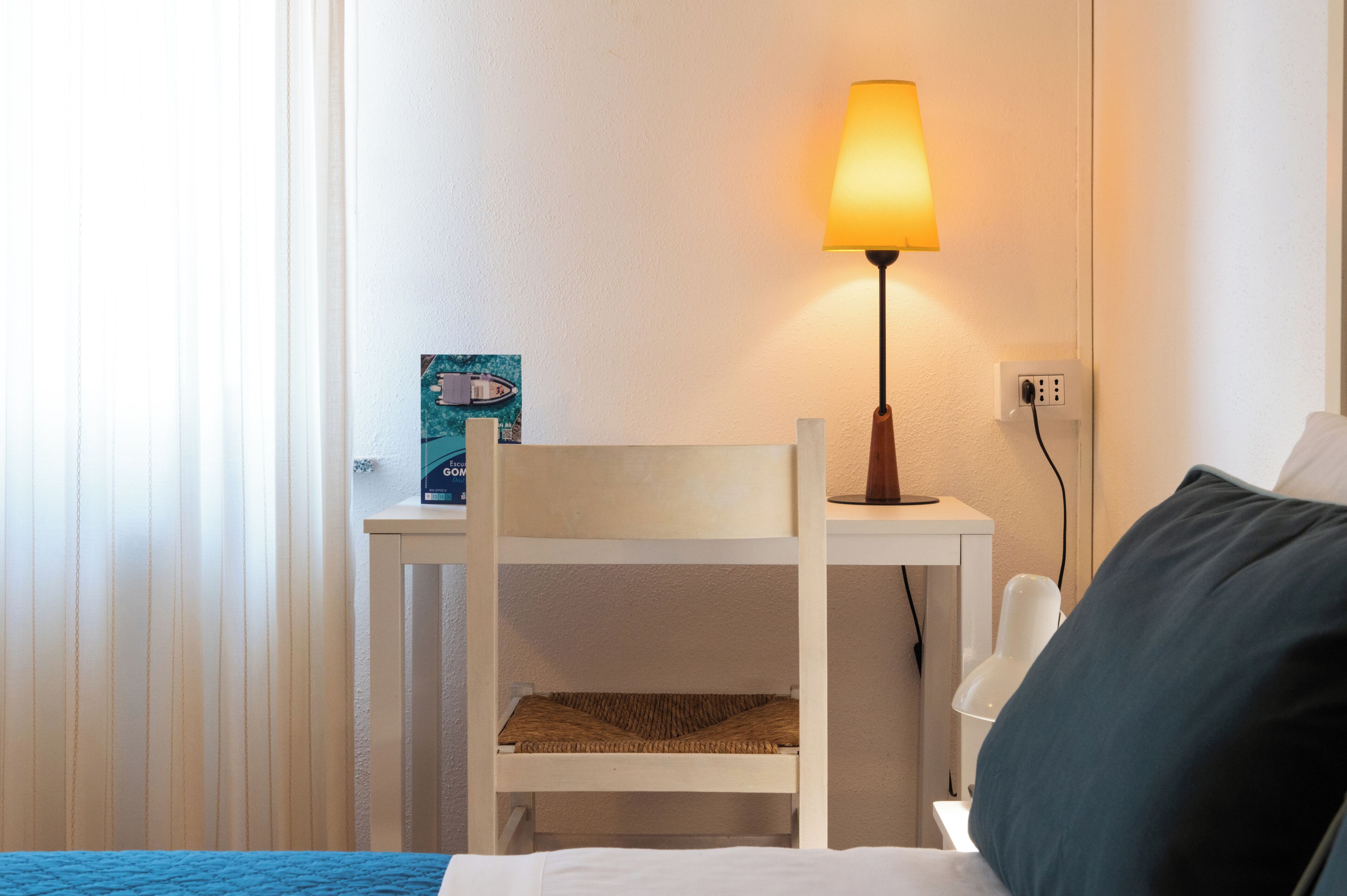 Quarto com cama, escrivaninha com luminária, cadeira e pequena mesa com porta-retrato.