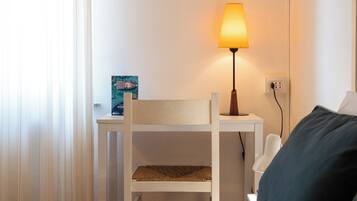 Quarto com cama, escrivaninha com luminária, cadeira e pequena mesa com porta-retrato.