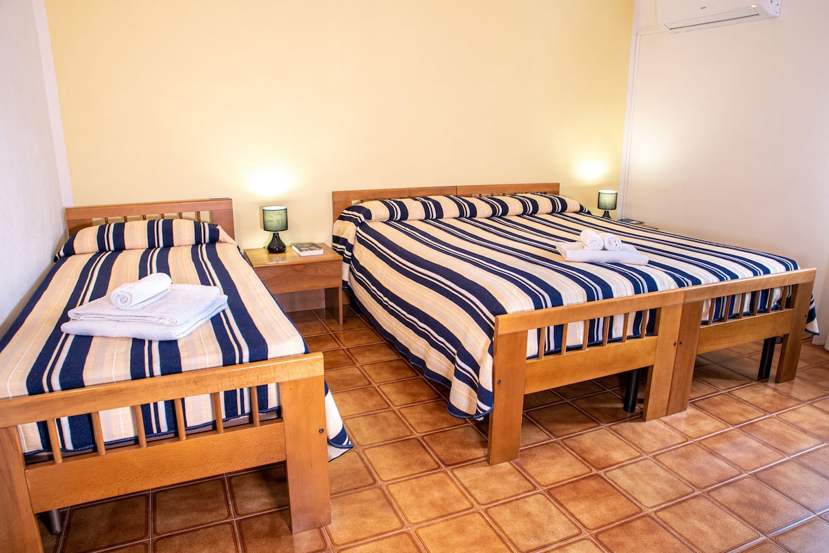 Camera d'albergo con due letti, ognuno con biancheria a righe e una testiera in legno. Sono presenti comodini con lampade e un condizionatore d'aria fissato al muro.