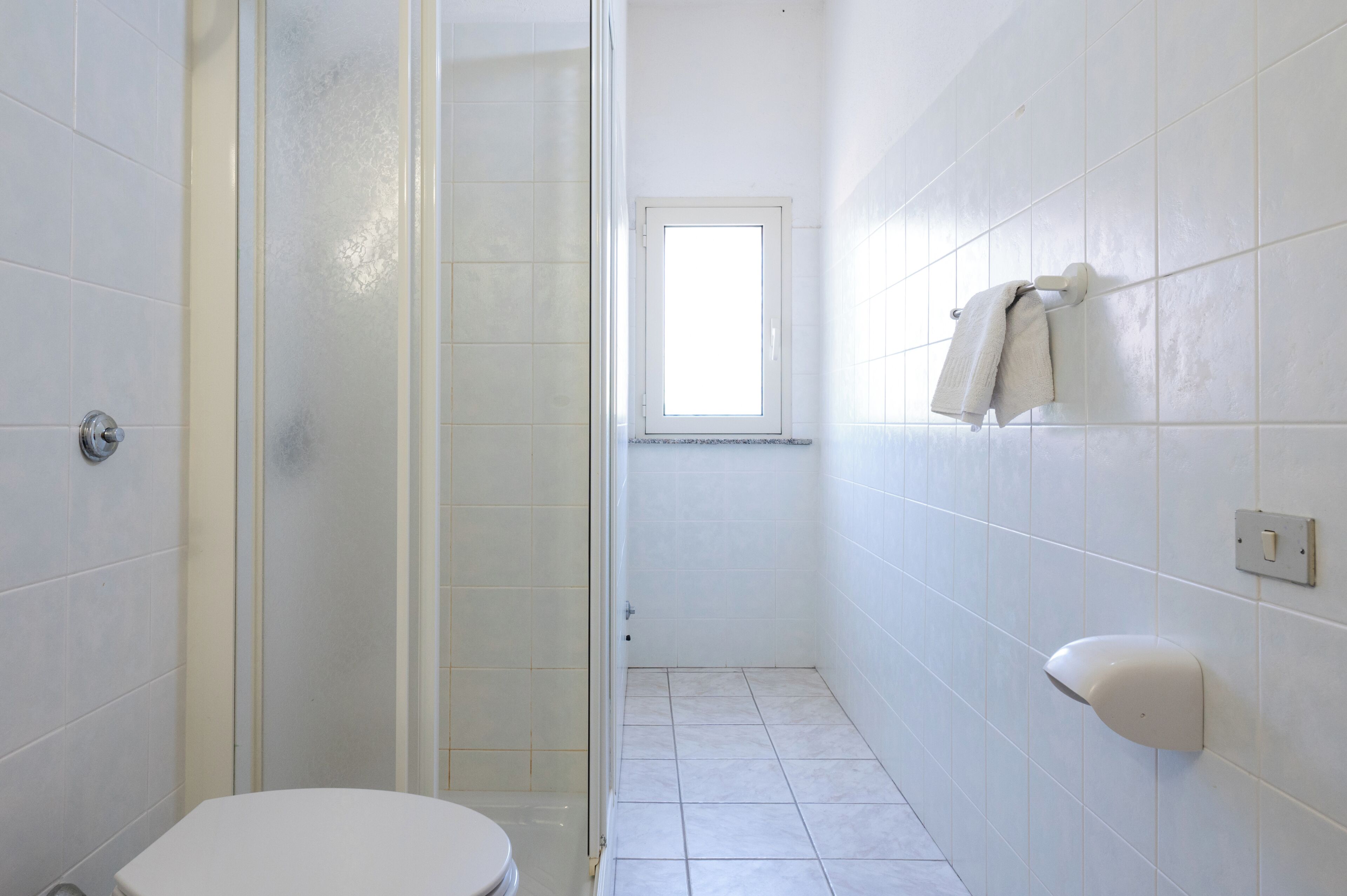 Une salle de bain avec des carreaux blancs, des toilettes, une douche avec une porte en verre, une fenêtre et un porte-serviettes.