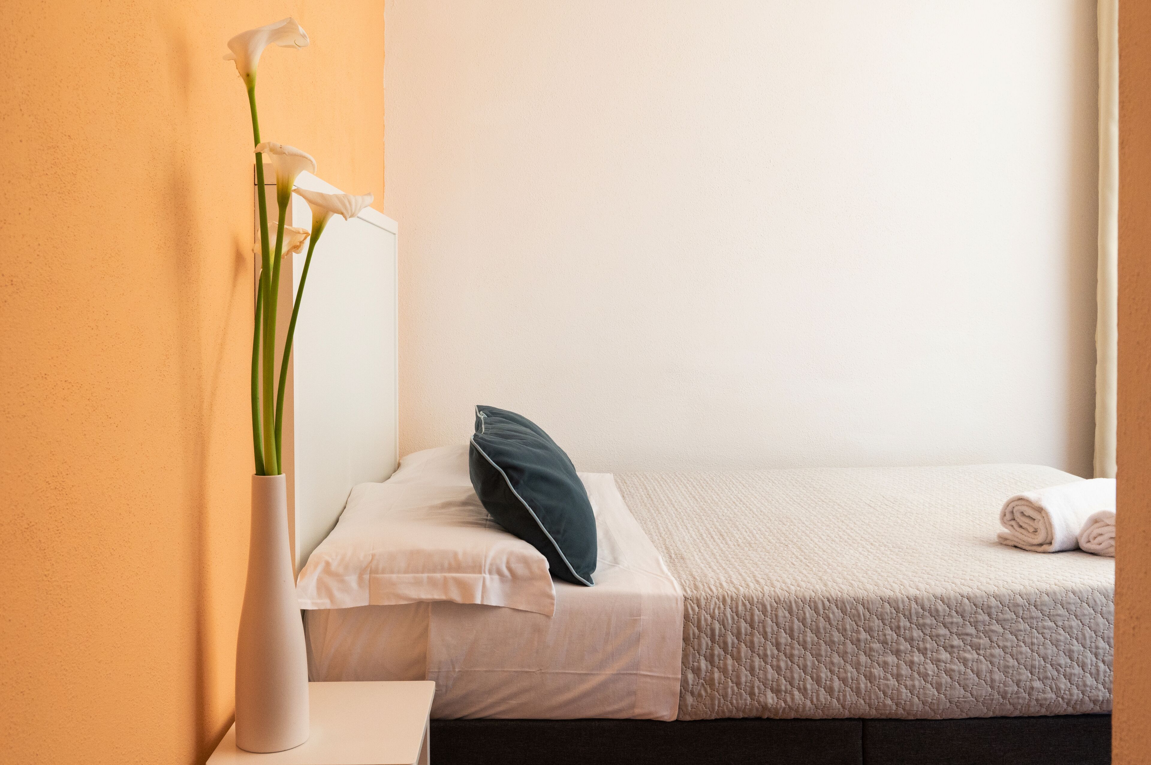 Una habitación de hotel con una cama, almohadas, un jarrón con flores y una pared.