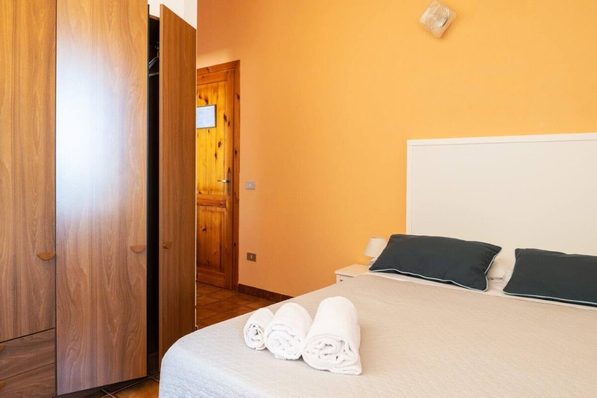 Una camera d'albergo con un'armadio in legno, un letto con biancheria bianca e due cuscini scuri, e un comodino con una lampada.