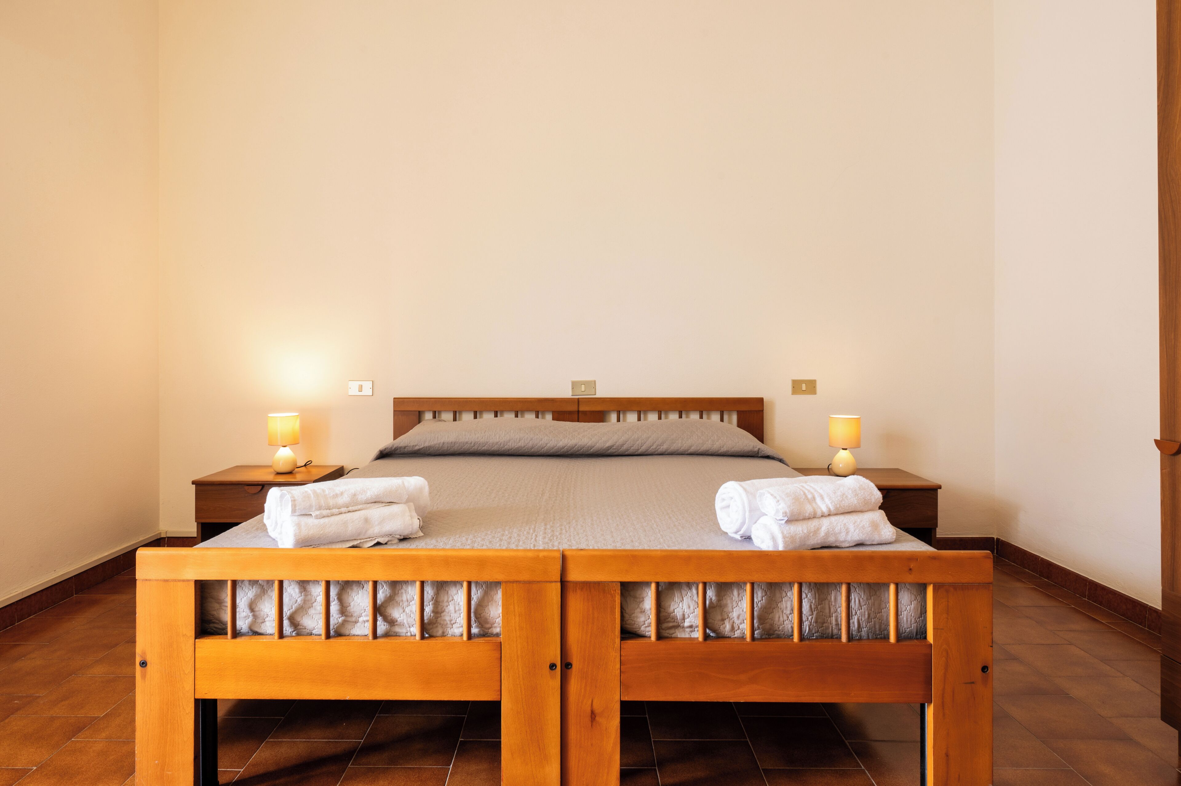 Ein Doppelbett mit Holz-Kopf- und Fußteil, zwei Nachttische mit Lampen sowie ein ordentlich bezogenes Bett mit Handtüchern.