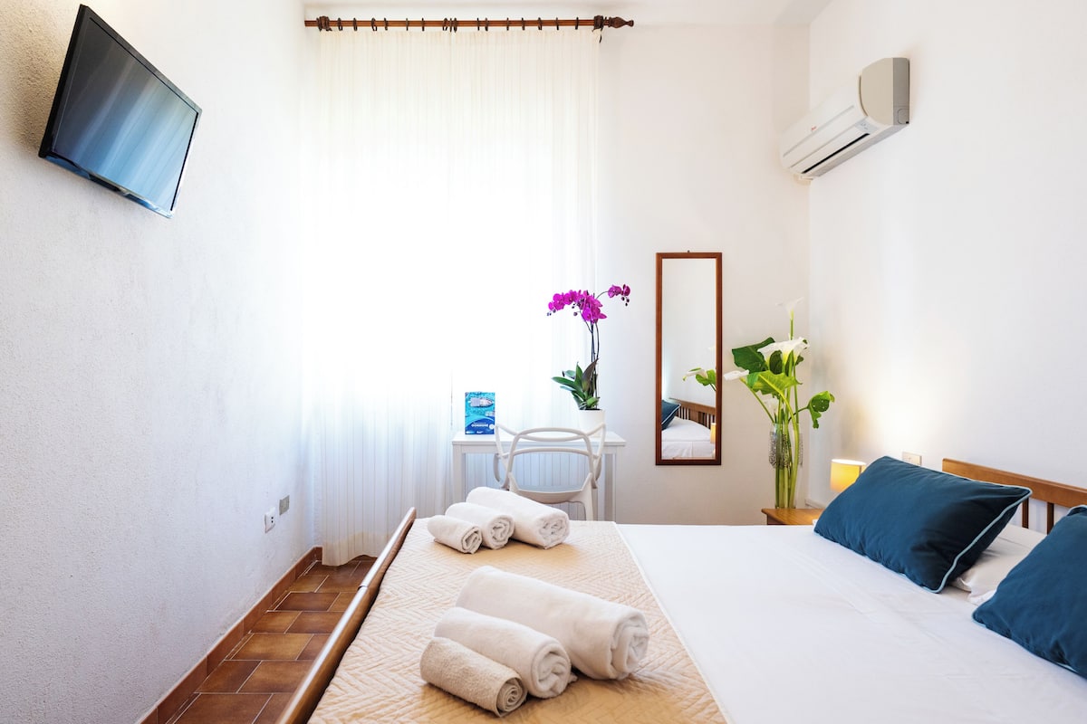 Una camera d'albergo con un letto, una scrivania, una sedia, una televisione, l'aria condizionata e una finestra con tende.