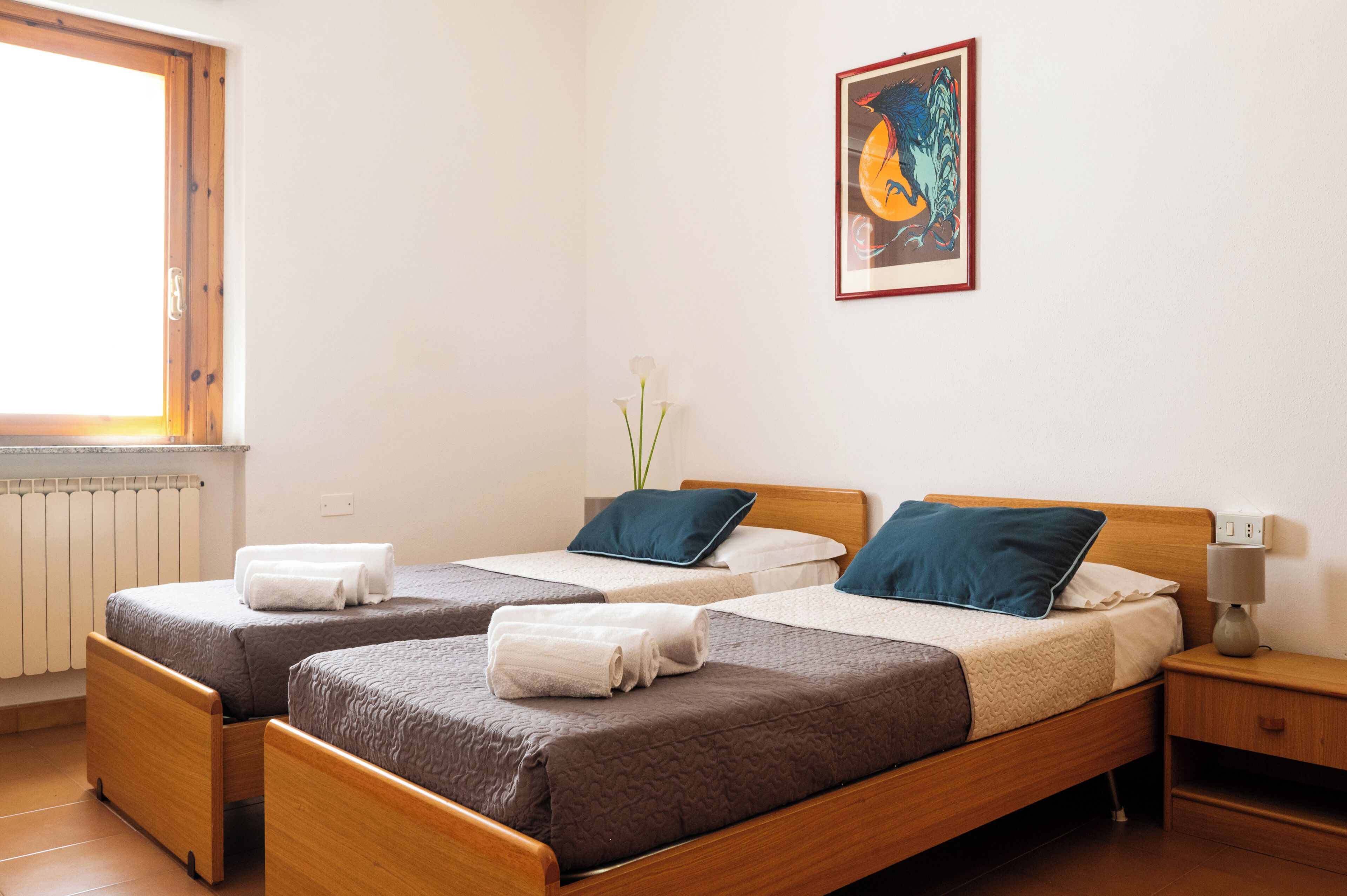 Habitació amb dos llits de fusta, tovalloles blanques, una obra d'art emmarcada, un ram de flors blanques en un vas i una taula de nit amb una làmpada.