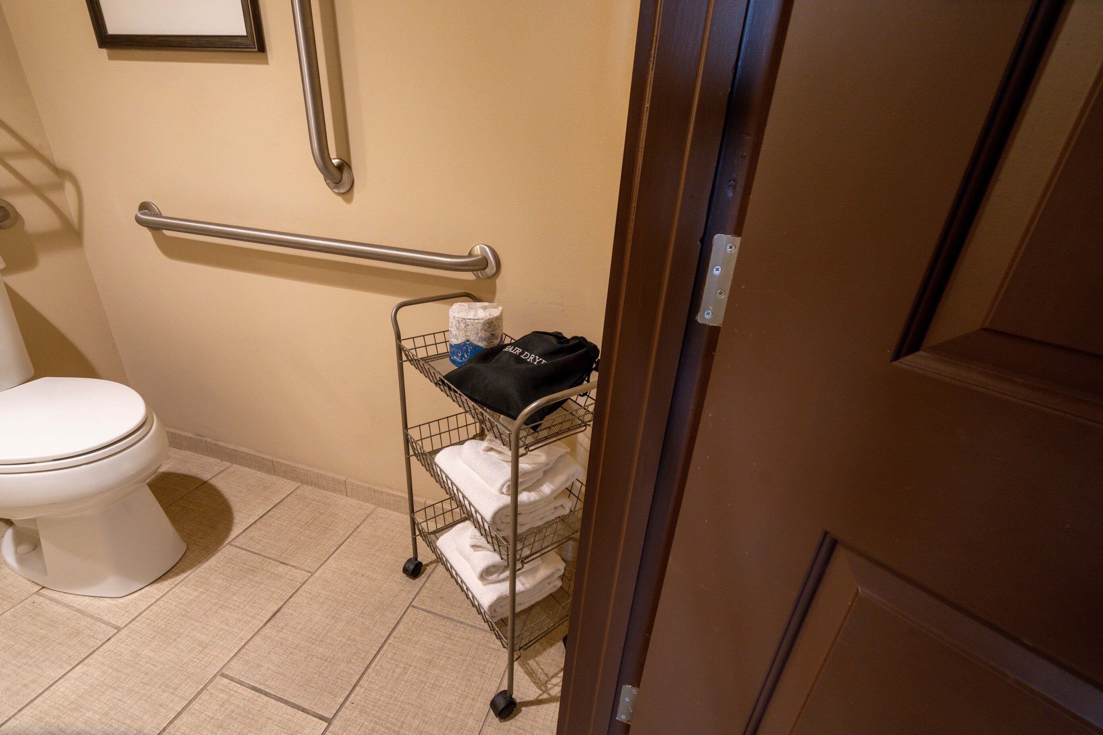 A bathroom with a toilet, a handrail, and a cart with towels and a bag.