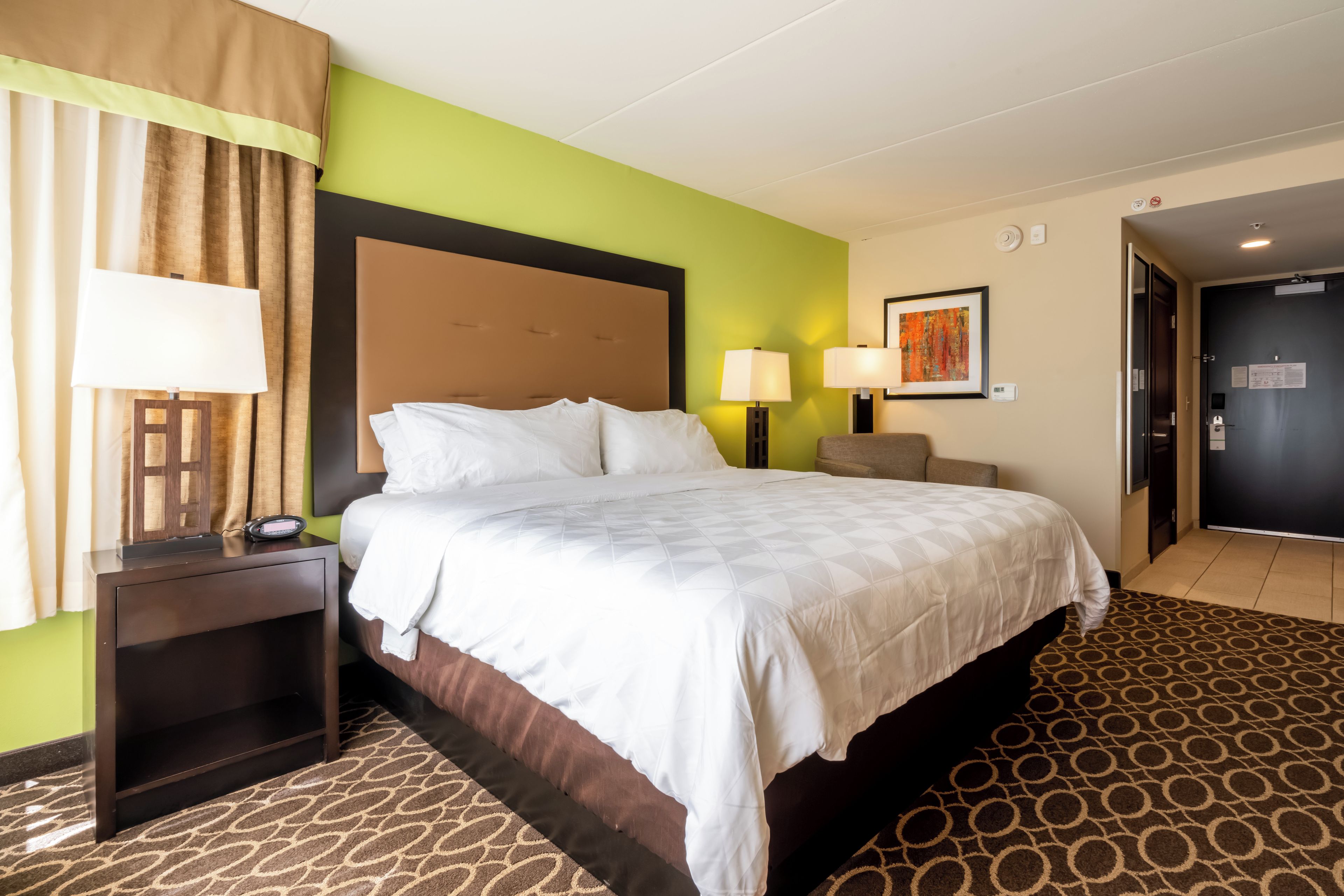 Een hotelkamer met een bed, nachtlampjes, een nachtkastje, een hoofdbord, een muur met een schilderij en een deur.