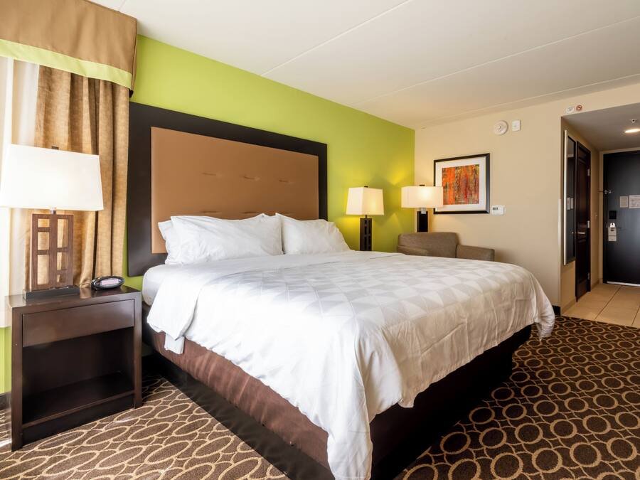 Een hotelkamer met een bed, nachtlampjes, een nachtkastje, een hoofdbord, een muur met een schilderij en een deur.