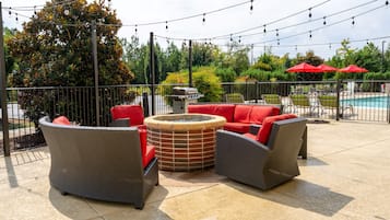 An outdoor seating area with wicker furniture and red cushions, a fire pit, and string lights.