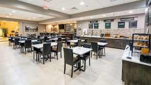 Una cafetería amplia con varias mesas y sillas, un mostrador con diversas opciones de comida, y un reloj en la pared.