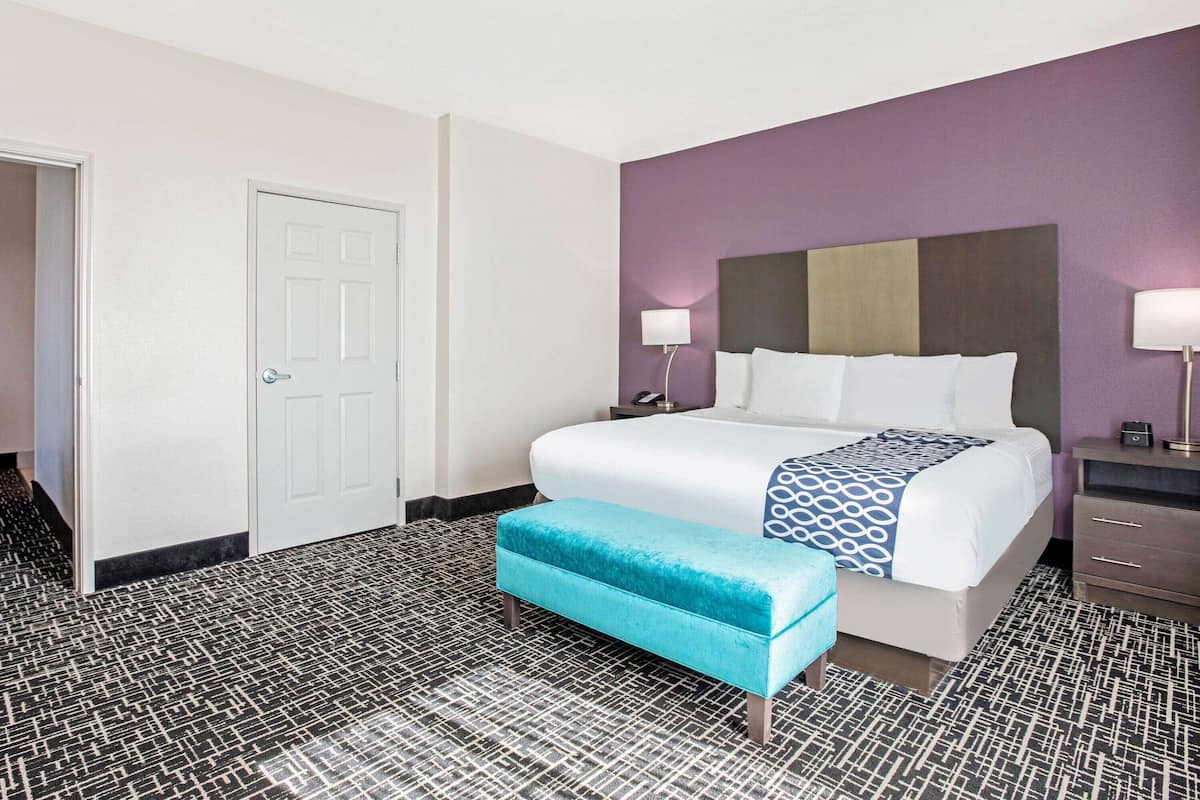 Une chambre d’hôtel avec un lit, une banquette, une table de chevet, une lampe et une porte.