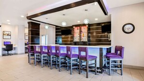 A modern bar area with a long counter and multiple purple bar stools. There is a television mounted on the wall and a clock above it. - La Quinta Inn & Suites by Wyndham Corpus Christi - Portland (Portland)