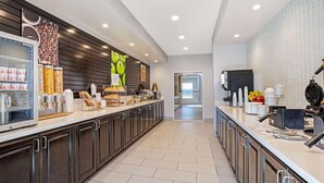 A modern kitchen area with a long counter, various food items, and a refrigerator. - La Quinta Inn & Suites by Wyndham Corpus Christi - Portland (Portland)