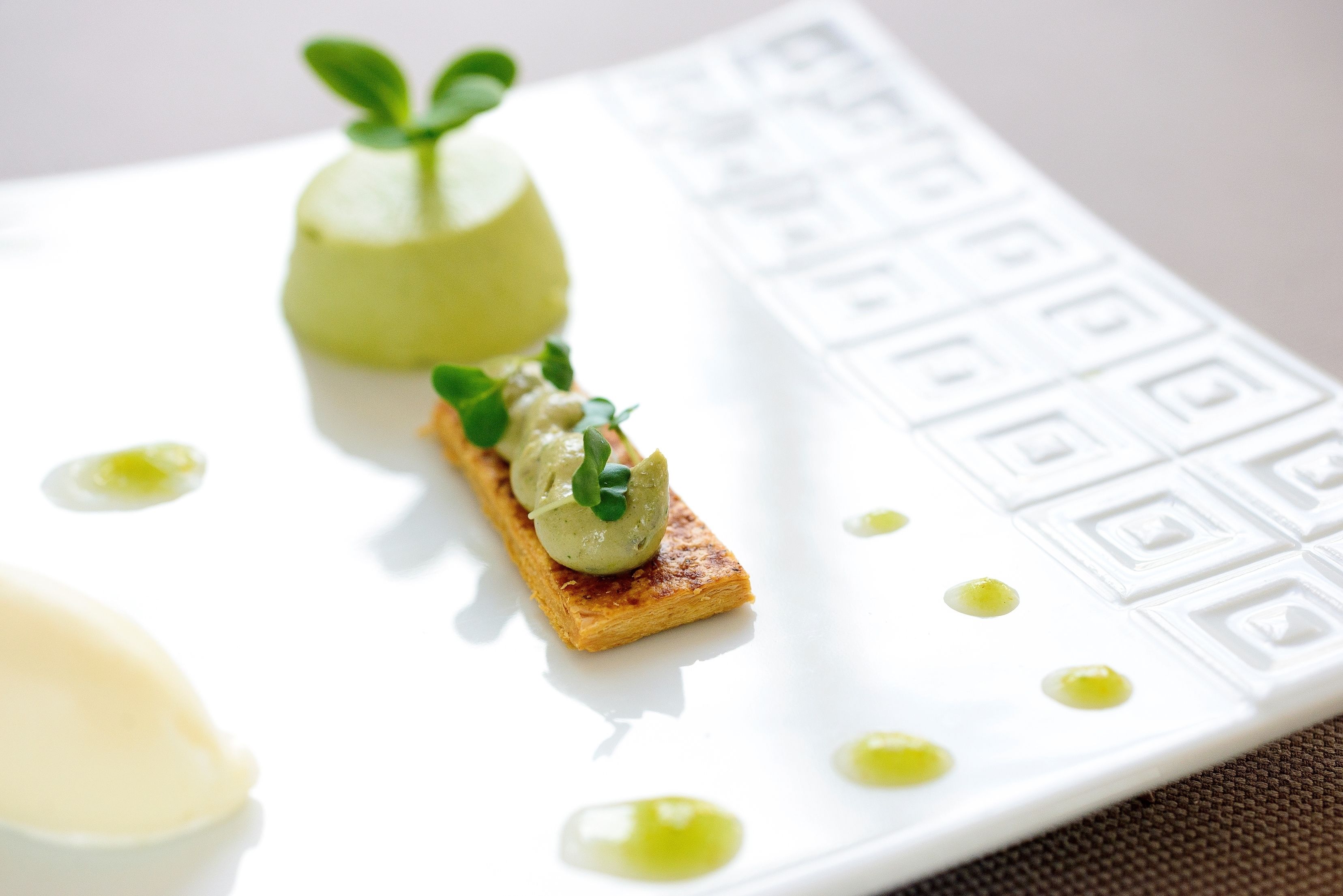 Un plato blanco con una rebanada de tostada cubierta de salsa verde y una pequeña guarnición verde, acompañado de algunas gotas de salsa amarilla sobre una superficie blanca con textura.