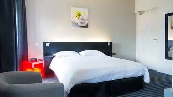 A hotel room with a bed, a bedside table, a chair, a wall-mounted artwork, and a door with a notice.
