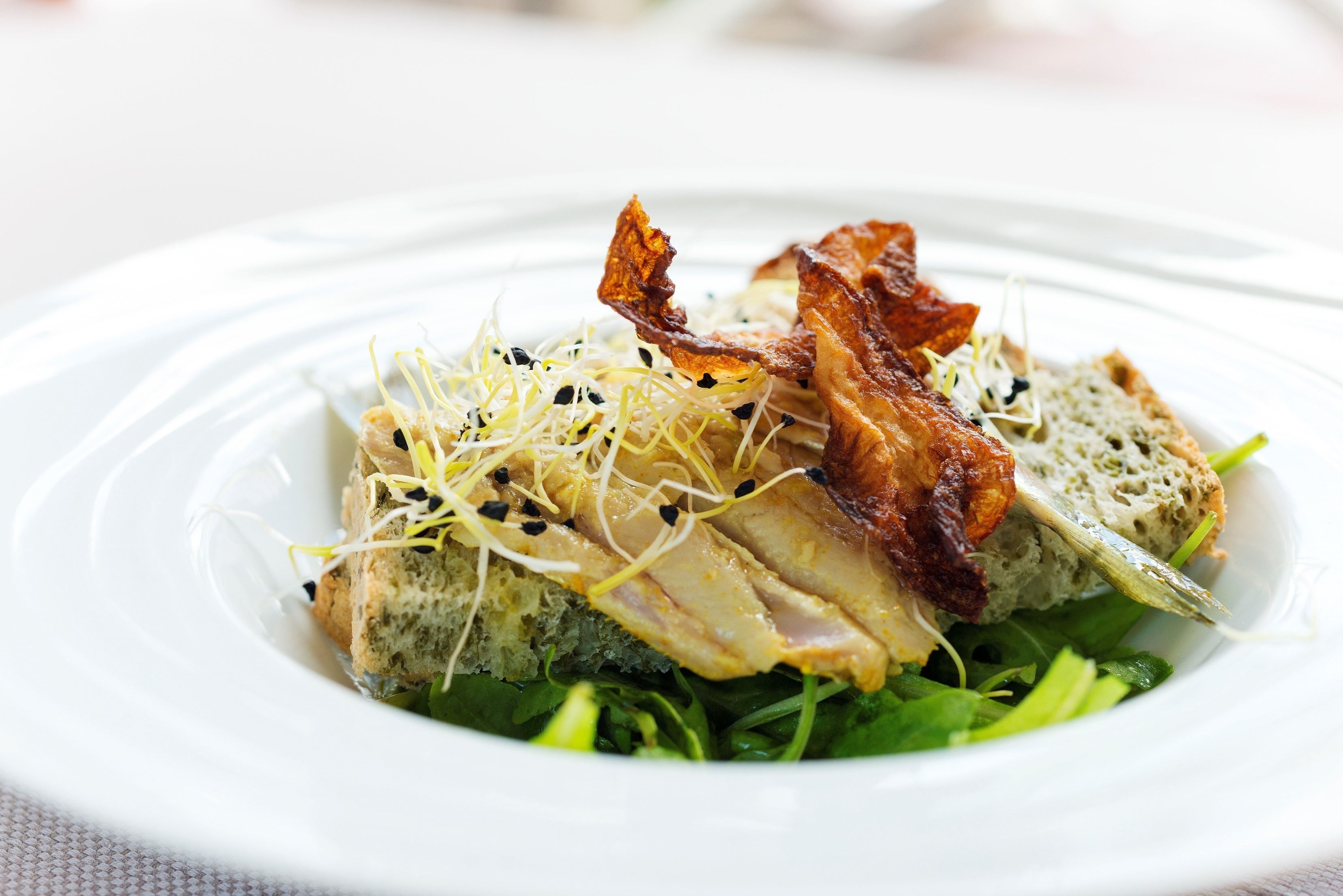 A white plate with a dish containing greens, a piece of bread, and crispy bacon.