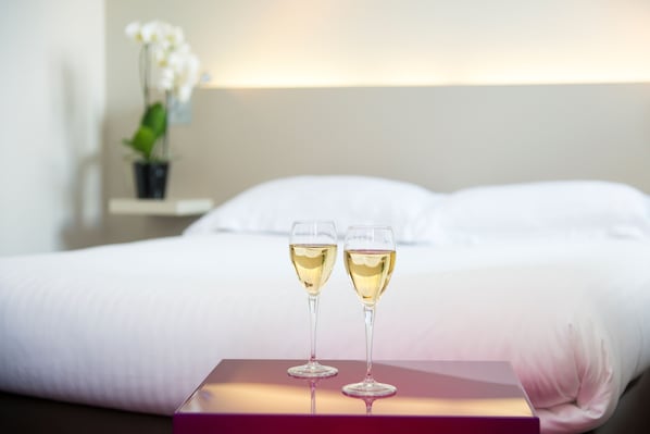 Two glasses of white wine on a table in a hotel room with a bed and a nightstand with a plant. - Le Germinal (Cesson-Sevigne)