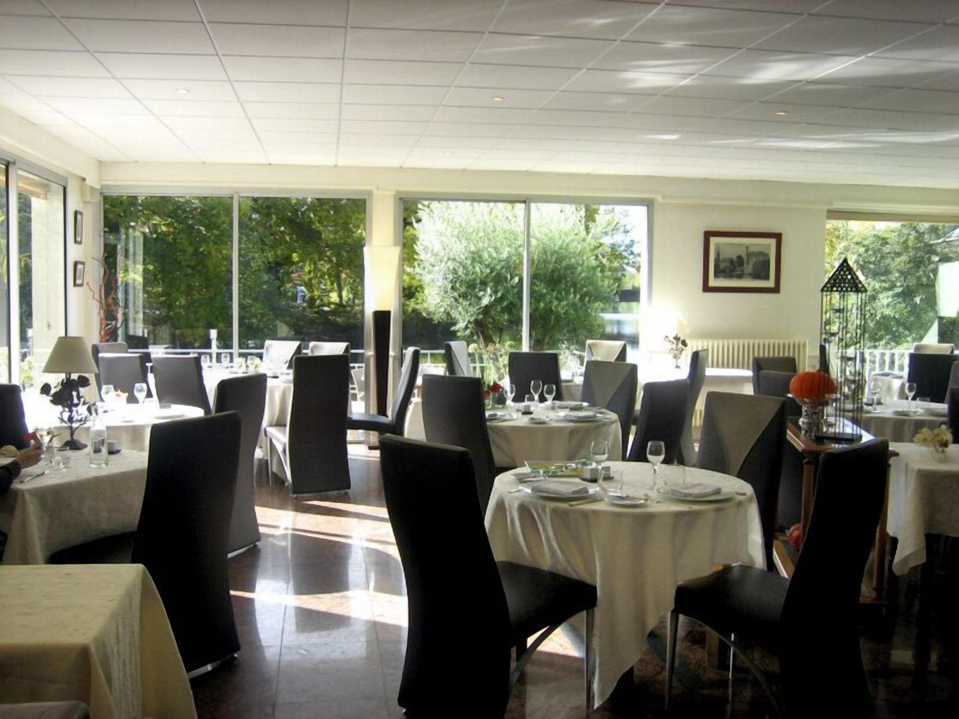 A restaurant with round tables, black chairs, and large windows offering a view of greenery.