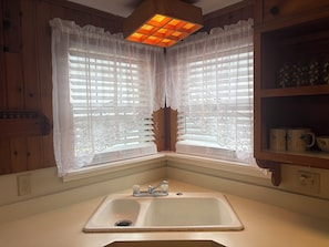A corner kitchen with a sink, wooden cabinets, and a window with lace curtains. - Nock Apartments (Ocean City)