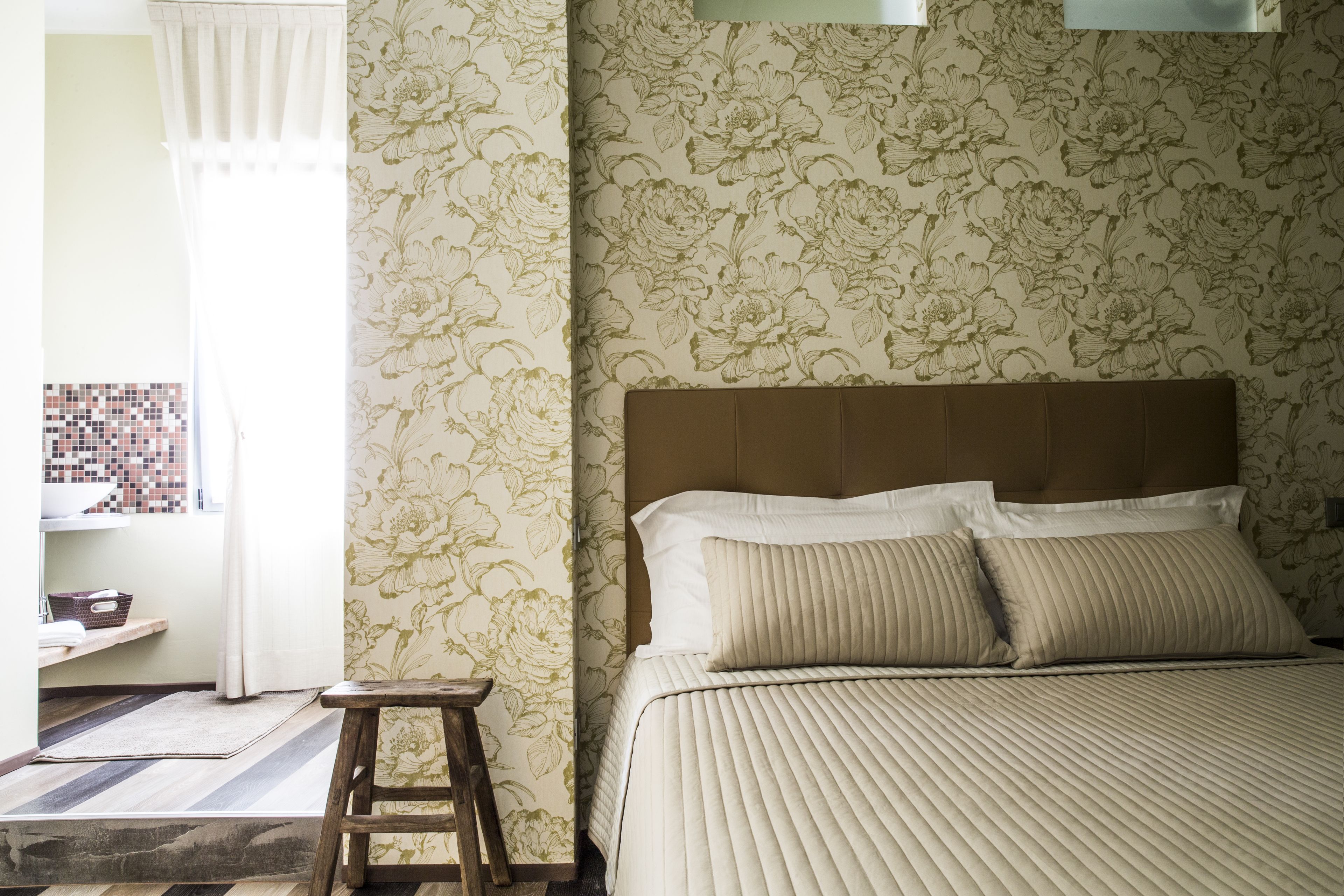 Un dormitorio con una cama, un taburete de madera, una ventana con cortinas y un papel tapiz floral en la pared.