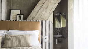 A bedroom with a large bed, a headboard, a bathroom with a sink and mirror, and a sloped ceiling with circular cutouts. - Serendipity Rooms (Milan)