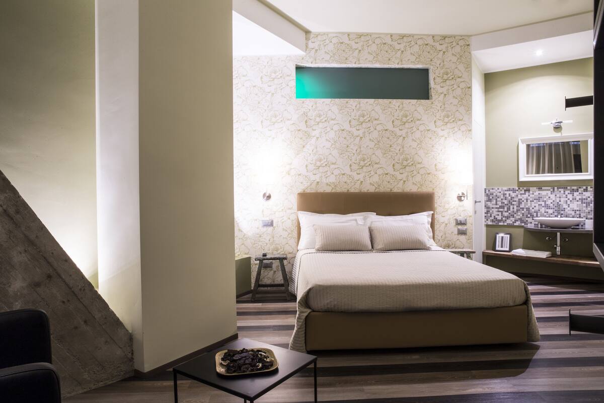 A modern bedroom with a large bed, a small table, a nightstand, and a bathroom with a sink and mirror.