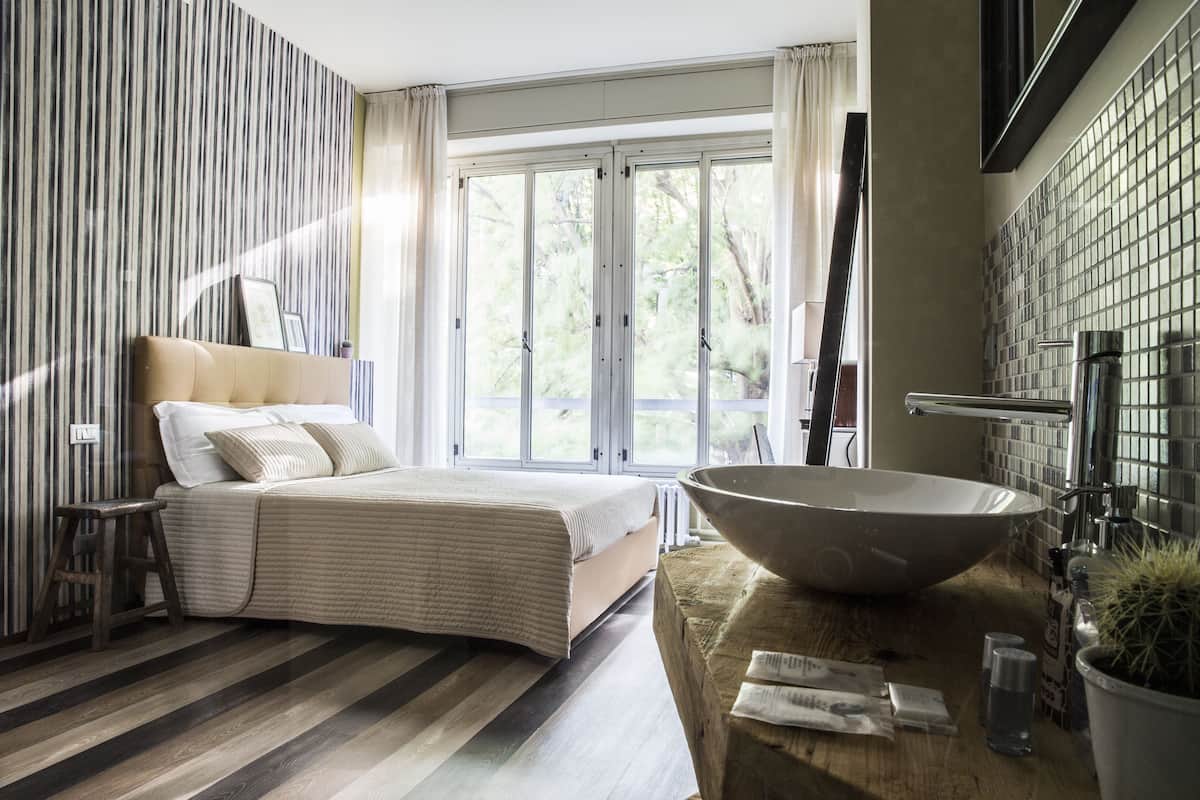 A bedroom with a large bed, a wooden stool, a large bowl sink, and a view of the outdoors.