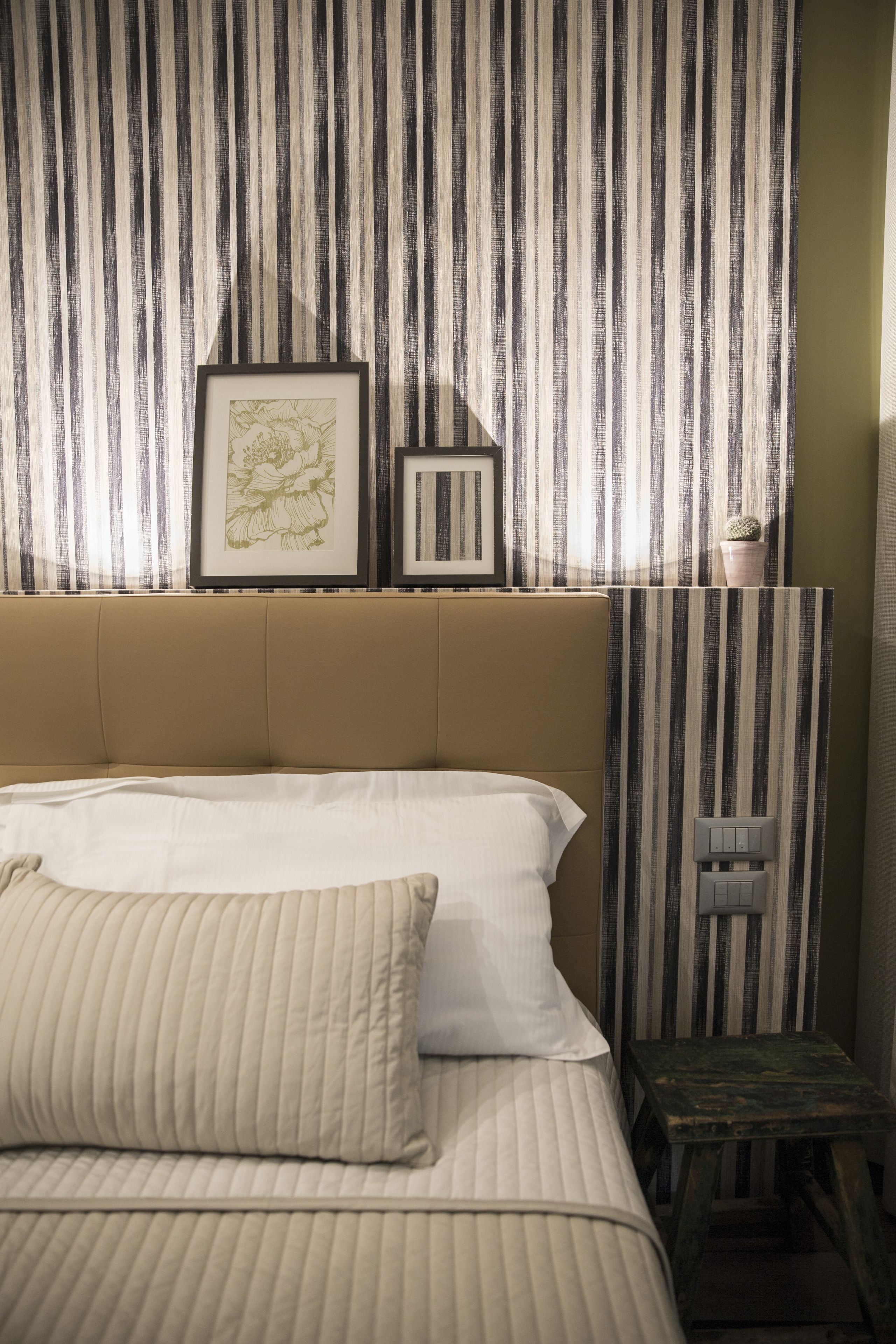 Un dormitorio con papel tapiz a rayas, una cama con ropa blanca, un cuadro enmarcado y un pequeño cactus en un estante.