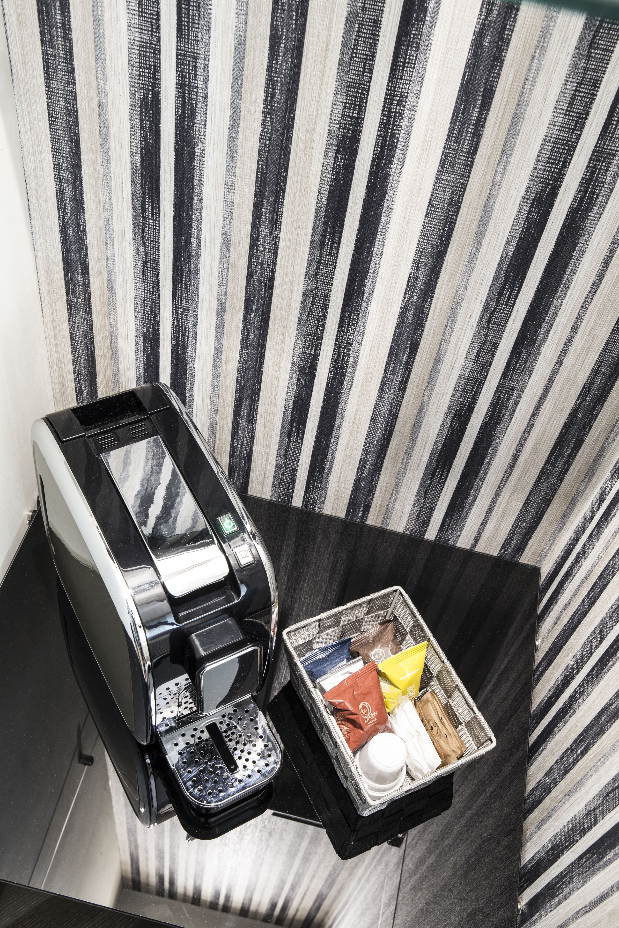Eine Kaffeemaschine und ein Tablett mit Kaffeekapseln und einer Tasse auf einem schwarzen Tisch, vor einer gestreiften Wand.