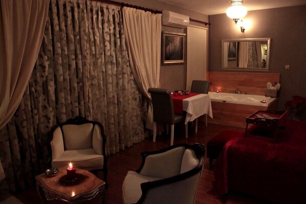 Una habitación tenuemente iluminada con una mesa de comedor, sillas, un sillón rojo, una mesita con una vela y un ventanal con cortinas.