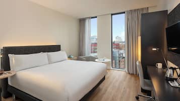 Quarto de hotel moderno com uma cama grande, uma escrivaninha, uma cadeira, uma TV e vista da cidade através de amplas janelas.