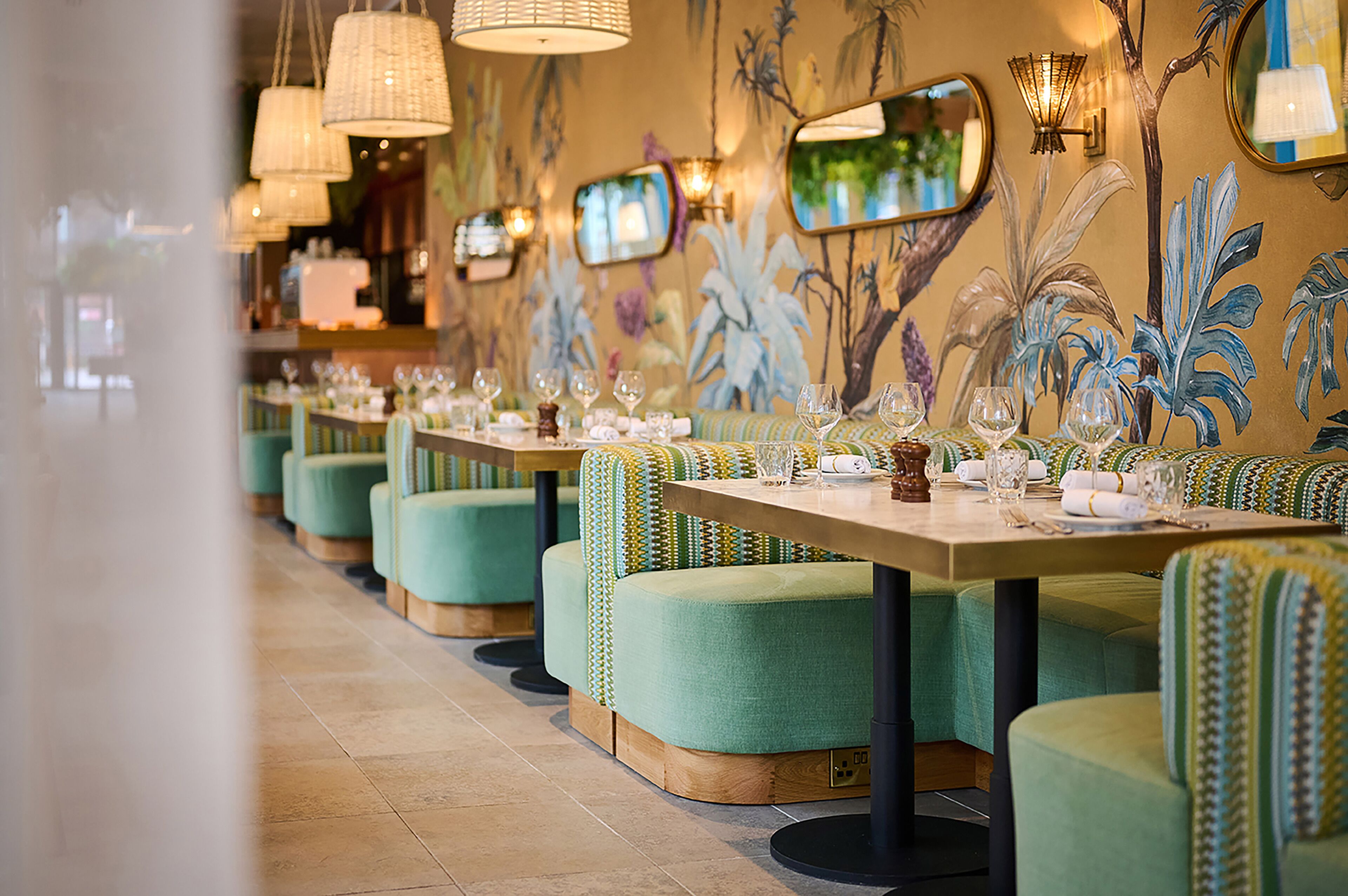A restaurant with green upholstered booths, a long table set with glasses and cutlery, and wall art depicting tropical foliage.