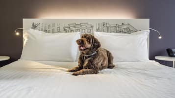 A dog sitting on a bed with white bedding and a headboard featuring a black and white cityscape drawing.