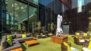 An outdoor seating area with a modern design, featuring a mix of sofas, chairs, and tables, surrounded by a well-maintained lawn and large windows offering views of the cityscape.